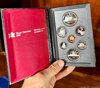 Open leather case showing seven proof coins, including a silver dollar with sailboat and a 50-cent piece, arranged in custom foam slots.