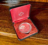 Coin in a red velvet-lined black presentation case with silver maple leaf emblem, showing the coin inside a protective capsule.
