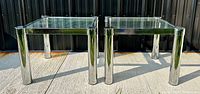 Pair of square mid-century modern end tables with chrome tubular legs and clear glass tops shown outdoors on concrete, highlighting the minimalist modern design.