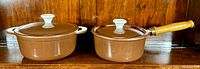 Image shows two brown cast iron cookware pieces on wooden surface: a larger pot with lid and handles on left, smaller saucepan with wooden handle and lid on right.