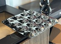 Photo of a square stainless steel fruit holder with a distinctive raised floral or abstract pattern on the surface. Mirror polished finish with reflections showing light wear.
