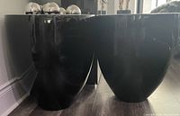Two glossy black ceramic planters side by side on wooden floor, reflecting surroundings.