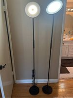 Two black Joofo floor lamps lit at different brightness and color temperature settings, showing round LED heads and tall slender poles on round bases.