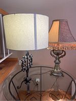 Two table lamps positioned on a round glass table: on left a metal base lamp with cylindrical cream shade, on right an ornate ceramic lamp with decorated shade and tassel fringe.