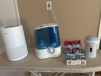 White Levoit air purifier, Vicks humidifier, small bedside humidifier, boxes of demineralization cartridges and mineral absorption pads on a table