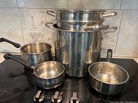 Set of stainless steel pots and pans arranged on stovetop including large and small pots, pans, and inserts