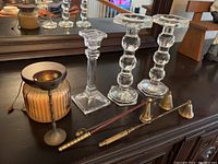 Photo of three glass candle holders, an orange wax candle in rippled glass jar, two candle extinguishers, and a small brass goblet-shaped item on dark wood surface.