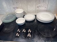 View showing all dishes together: stacks of five clear glass plates, five white plates, five white saucers, and five white bowls on kitchen stove and counter.