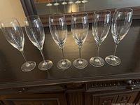 Six clear stemmed wine glasses arranged in a row on a dark wood surface, showing overall shape and clarity.