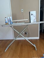 Full-size folding ironing board with padded beige cover, metal legs, standing open on wooden floor, with Vela garment steamer box and cans of starch on board surface.