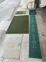 Overview of the golf practice set showing the full swing mat, putting mat, chipping mat, and chipping basket laid out on a concrete floor near a garage.