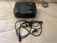 InFocus projector with remote control placed on top and VGA and power cables in front, alongside measuring tape for scale.