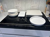 Set of white dishes on stovetop including seven large square plates, two white bowls stacked, and one rectangular platter