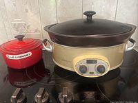 Photo showing both Le Creuset red pot and Versa Ware Pro slow cooker on stove, with lids on.
