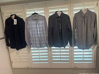 Four men's long sleeve shirts hanging on hangers in front of white shutters. Shirts are black, gray and blue checkered, black dotted, and gray with buttoned collar.