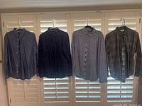 Four men's button-up shirts hanging on white hangers in front of window shutters: dark gray, navy checked, light gray, and dark green plaid.