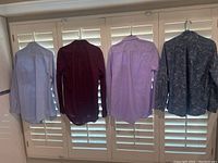 Back view of four long-sleeve men's button-up shirts in light blue, dark maroon, lavender, and dark gray paisley pattern hanging on hangers in front of window shutters.