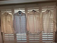 Four white men's dress shirts hung on hangers, displayed on window blinds. Each shirt is long-sleeved with front buttons and chest pockets visible on some. Shirts appear used and vary slightly in style and fabric thickness.