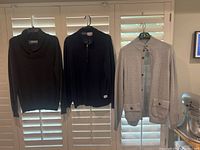 Front view of three sweaters hanging on hangers in front of window blinds - black sweater, black dotted sweater, gray button-up collar sweater