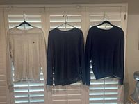 Three men’s cotton sweaters on hangers in front of window shutters: one light gray with logo, two dark blue without logos.