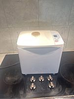 Front and top view of Panasonic bread maker showing white body, round viewing window on top, and control panel with buttons and digital display.