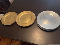 Three gold plate chargers with beaded rims, three gold plate chargers with grooved rims, and eight silver plate chargers with smooth rims, arranged on a table.