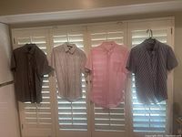 Four short sleeve dress shirts hanging on hangers against white plantation shutters, showing their full shirt form and colors.