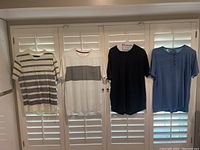 Four collarless short sleeve shirts hanging on hangers, left to right: white with stripes, white with chest band, black, grayish-blue with buttons.