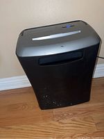 Front view of black and gray Staples document shredder placed on wood floor, showing wear and scuffs.