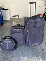 Photo showing three $ Tag luggage pieces in dark color with all handles extended, large, medium luggage suitcases with wheels and small carry-on bag.