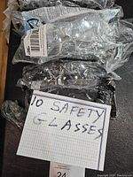 Top-down photo showing 10 new safety glasses individually sealed in plastic bags, some with visible labels and barcodes, positioned on a dark surface beside a note reading '10 SAFETY GLASSES'.