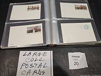Photo shows an open black album with postal cards in protective sleeves, handwritten note 'LARGE COLL POSTAL CARDS' visible.