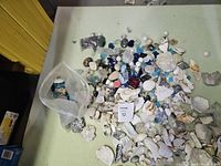 Wide view showing a large collection of mixed rocks, shells, coral pieces, and colored glass spread on a flat surface with a plastic bag containing some smaller items.