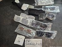 Photo showing entire lot of 10 sunglasses mostly wrapped in plastic or inside pouches on a dark surface; includes various frame styles and colors with dark-tinted lenses.