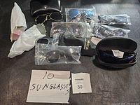 Lot of 10 new sunglasses displayed with some in packaging and several black cases and pouches included.