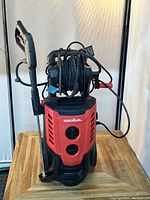 Front view of Benchmark pressure washer showing red and black body, control knobs, attached wand, coiled hose on reel, and electrical cord.