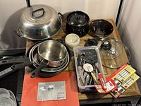 Photo showing entire set of culinary items laid out on a wooden surface, including pots, pans, utensils, kitchen knife set in packaging, and lids.