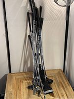 Photo showing multiple metal golf clubs standing on a wooden surface with black grips and steel shafts, various club heads visible.