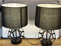 Set of two table lamps with black geometric metal bases and black fabric cylindrical shades. Shown side by side with cords visible, photographed against plain background.