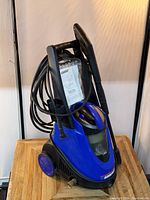 Blue Simoniz electric pressure washer with hose and attachments wrapped on handle, placed on a wooden surface, front and side views shown.