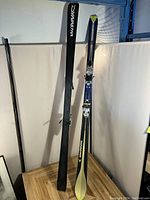 Dynastar ski with binding and accompanying ski pole, shown standing upright on wooden surface against white backdrop.