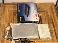 Sony Blu-ray Disc/DVD player components including the main unit wrapped in protective material, cables (power and A/V), and instruction manual placed on wooden surface with the product box behind.