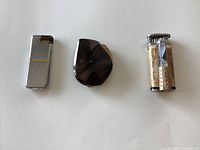 Photo showing three cigarette lighters: silver rectangular Saffa, dark glossy Flaminar, and orange-tan USA cigar lighter.