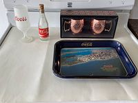 Photo showing all lot items on white stove surface: two copper mugs in box, Coca-Cola tray, Coors glass, Coca-Cola bottle