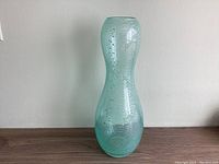 Full view of tall light blue glass vase with bubble pattern on wood surface against wall