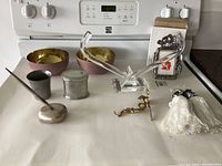 Brass coat hanger, acrylic hanger, two decorative bowls with gold interior, pewter cups, desk pen with metal base, new frame, and package containing brass hardware seen on white surface.