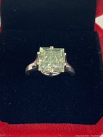 Front view of silver ring with a large square green Moissanite stone in the center, placed in a black velvet box.