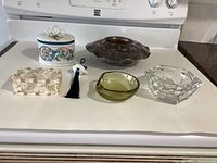 Full lot on kitchen counter showing Murano glass ashtray, porcelain candy dish with floral decoration, clear faceted glass dish, two decorative boxes, metal bowl, and dolphin figurine with tassel.