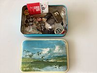 Photo of the open rectangular tin box with painted lid, showing various pins and medals inside.