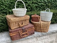 Overview of eight wicker baskets and suitcases stacked outdoors on a stone ledge. The basket colors are natural light brown, dark brown, and white. Different sizes and shapes are shown.